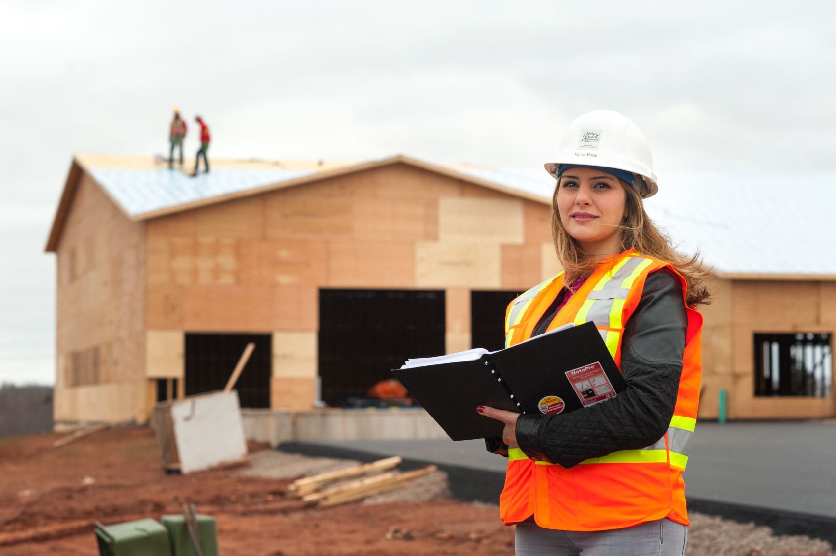 A construction worker in an orange safety vest and white hard hat holds a notebook while standing in front of a partially built wooden house, with two workers visible on the roof.
