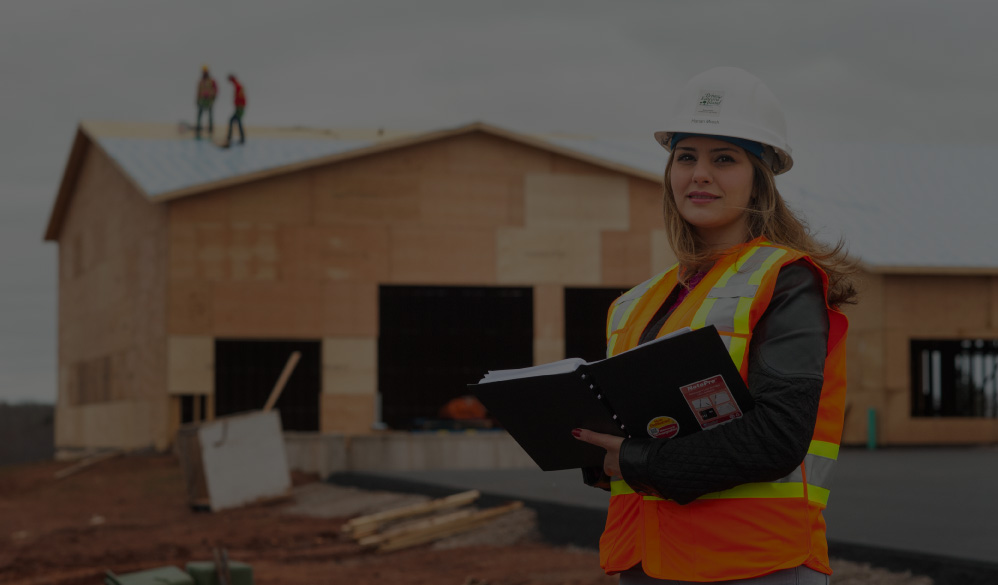 A construction worker in an orange safety vest and white hard hat holds a notebook while standing in front of a partially built wooden house, with two workers visible on the roof.