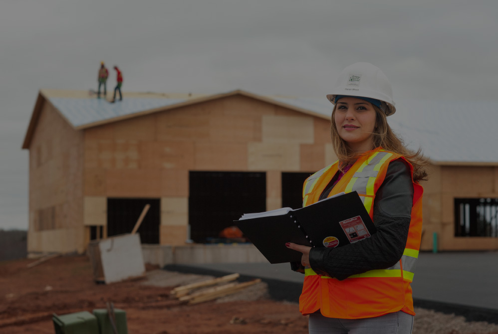A construction worker in an orange safety vest and white hard hat holds a notebook while standing in front of a partially built wooden house, with two workers visible on the roof.